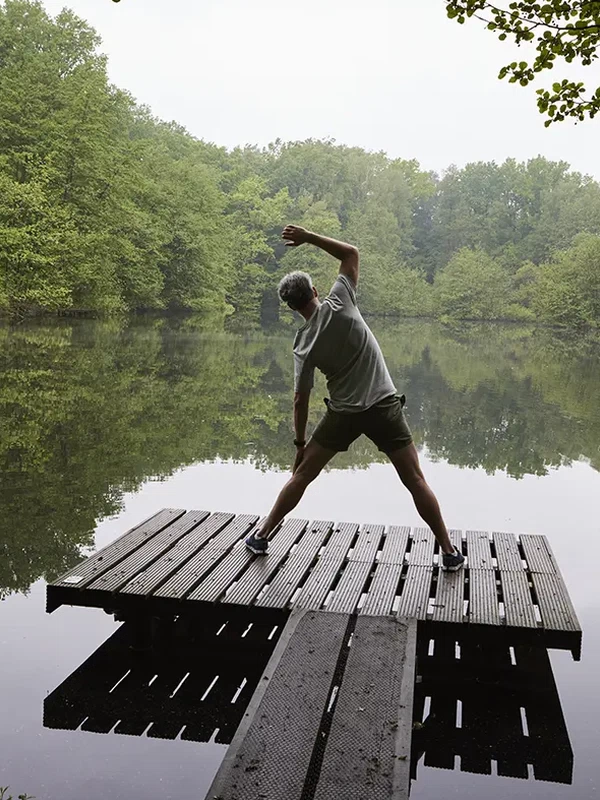 Eine Person macht Morgenstretching auf einem hölzernen Steg an einem ruhigen See, umgeben von dichtem Wald und Natur.