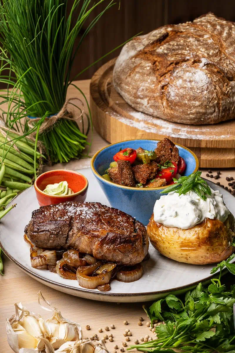 Saftiges Rumpsteak auf karamellisierten Zwiebeln, einer Ofenkartoffel mit Kräuterquark und frischem Bauernbrot, umgeben von Kräutern und Knoblauch.